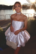 A woman wearing a strapless white satin mini dress with a flared skirt, holding a beige and white checkered handbag, set against an outdoor waterfront background at sunset.